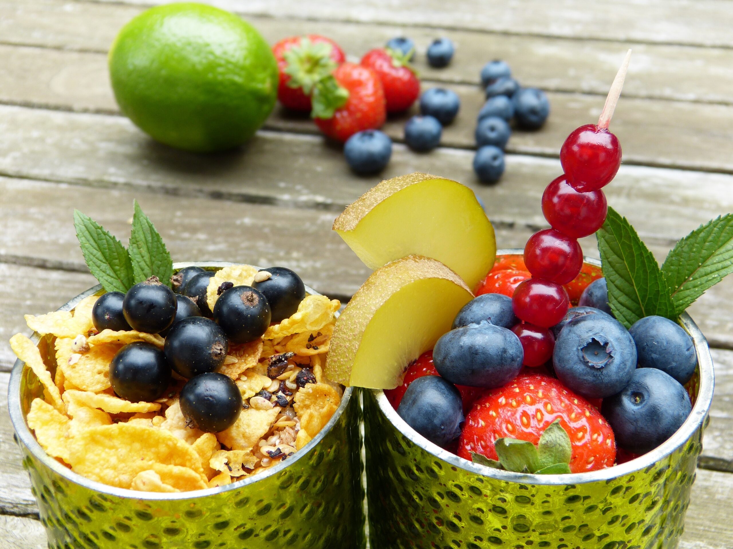 Zwei Gläser mit frischen Früchten, Müsli und Minzblättern, dekoriert mit Beeren, einer Limette und Erdbeeren im Hintergrund für blog Nahrungsergänzungsmittel